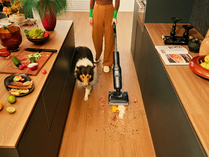 Жінка прибирає кухню ручним пилососом, а поруч із нею гуляє собака.
