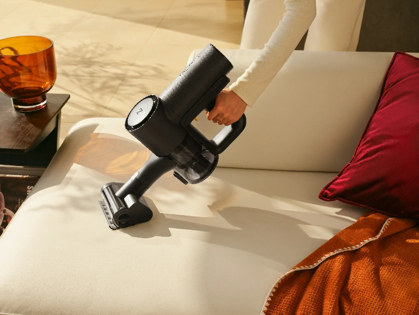 Жінка чистить оббивку дивана ручним пилососом.
