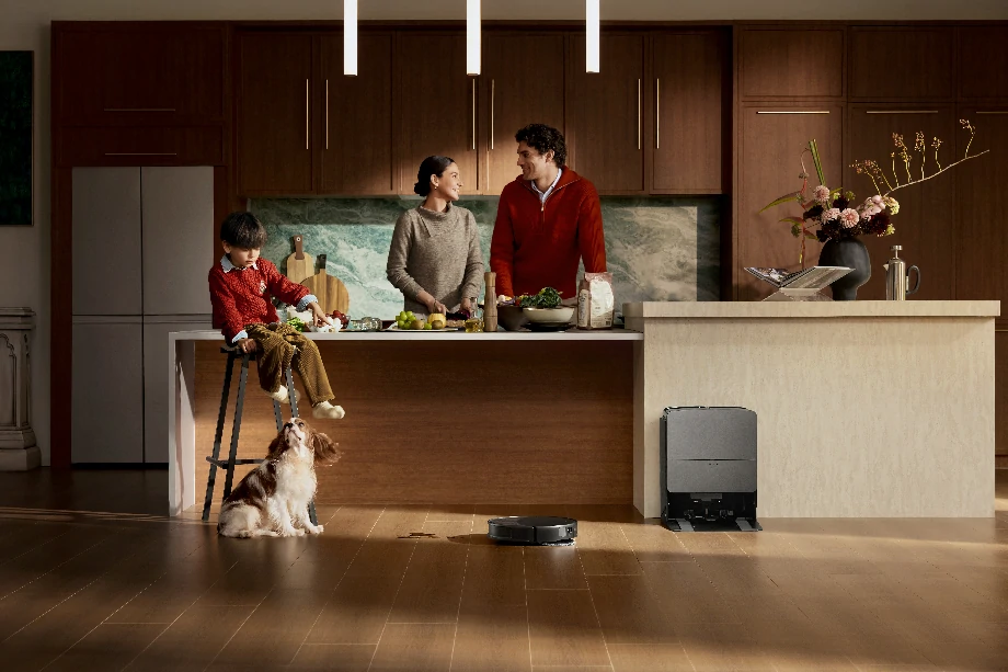 Пара готує їжу на кухні, хлопчик сидить у дитячому стільчику, а поруч із ним — собака. Перед ними — чорний робот-прибиральник.