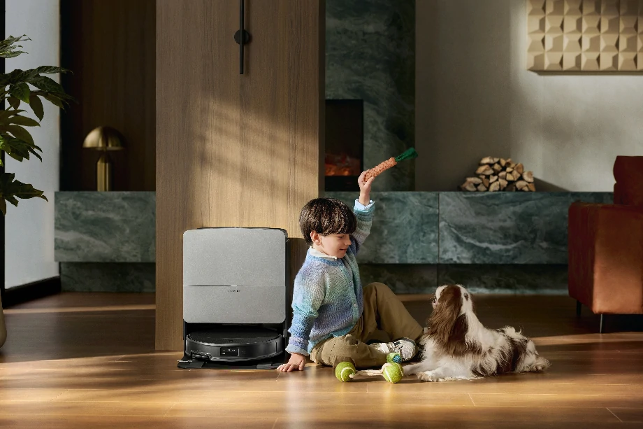 Хлопчик грається на підлозі вітальні зі своїм собакою, а поруч стоїть робот-прибиральник із док-станцією.