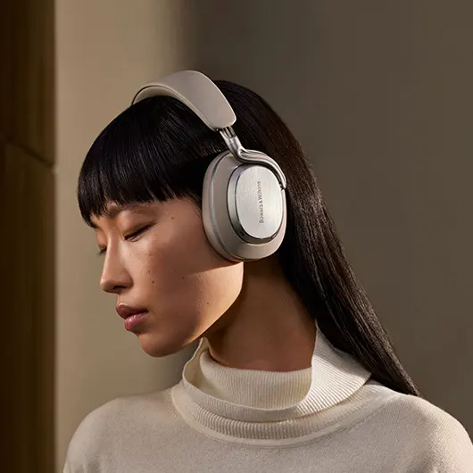 Жінка дивиться ліворуч і в навушниках Bowers & Wilkins Px8 S2 кольору «Теплий камінь».