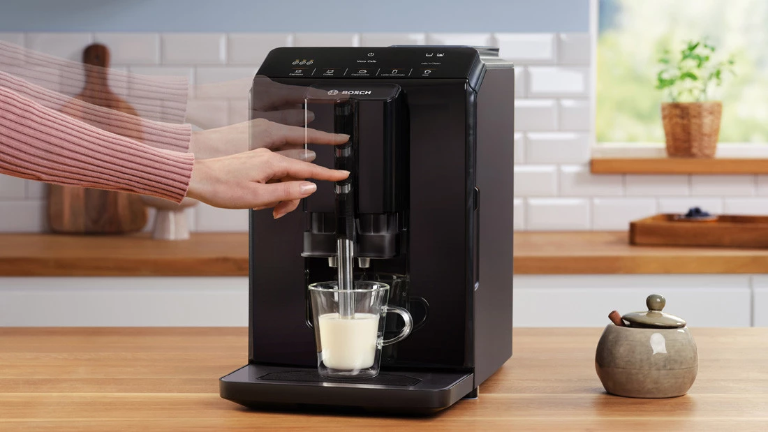 Кавоварка зі склянкою молока. Пальцем рухається насадка для спінювання молока до склянки.