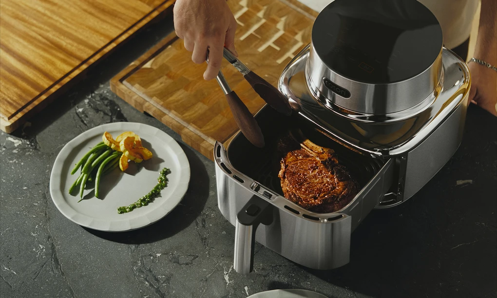 Рука, яка щипцями виймає шматок приготованого м'яса з відкритого кошика фритюрниці Cosori.