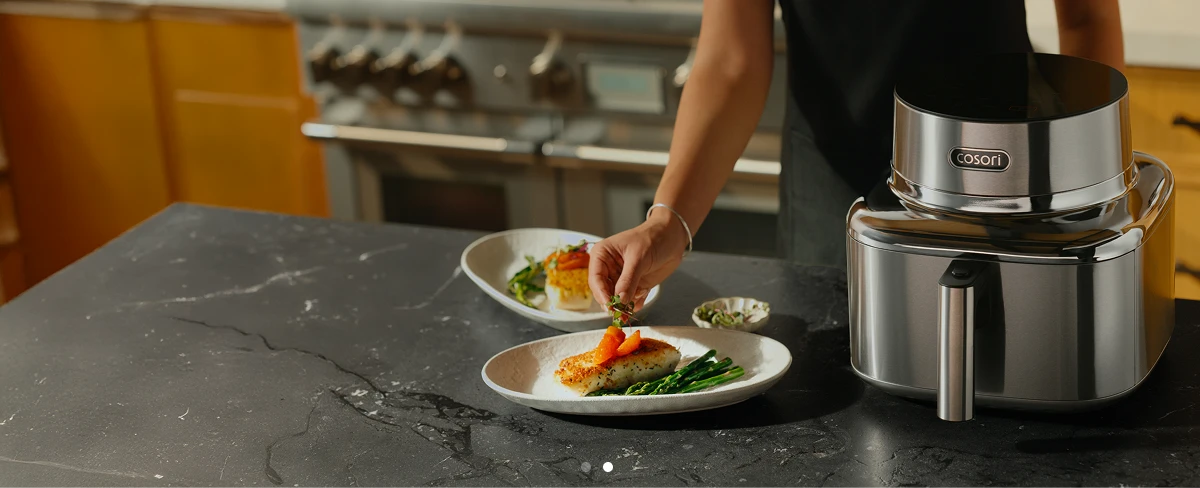Жінка, яка прикрашає готову рибну страву, стоїть поруч зі срібною аерофритюрницею Cosori Iconic Single.