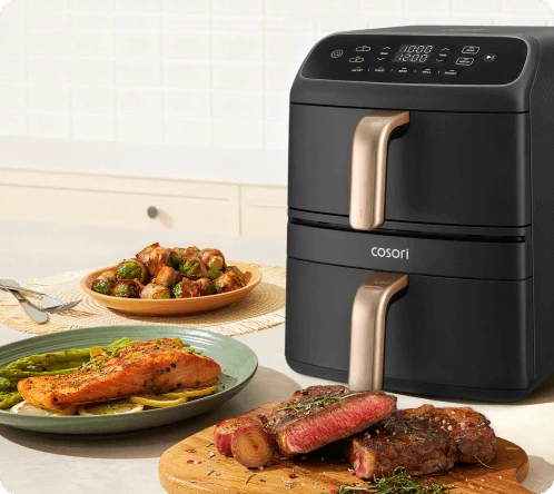 Фритюрниця Cosori Turbo Tower Compact стоїть на кухонному столі, оточена різними стравами, зокрема лососем та стейком.