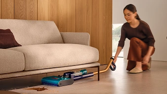 Жінка тримає в руці швабру, складену паралельно підлозі, і прибирає простір під диваном.