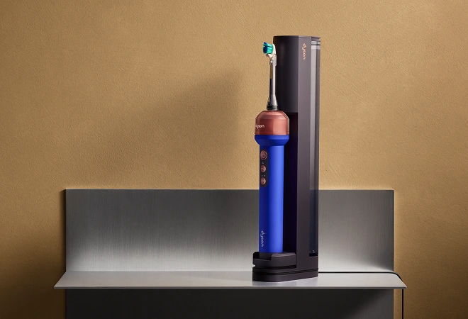Синя електрична зубна щітка Dyson CameraJet стоїть у фіолетовій док-станції на сірій стільниці.