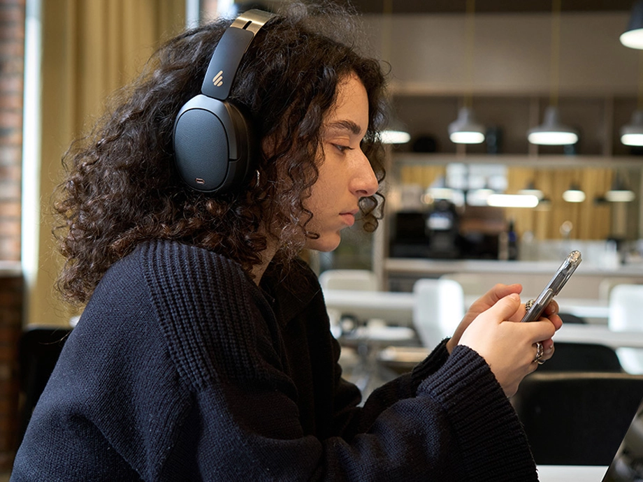 Жінка з кучерявим волоссям у темних навушниках-накладках тримає телефон у кафе