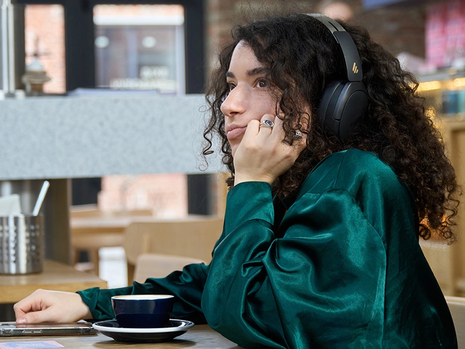 Молода жінка з кучерявим волоссям у темних навушниках-накладках тримає голову на руці за столом із чашкою.