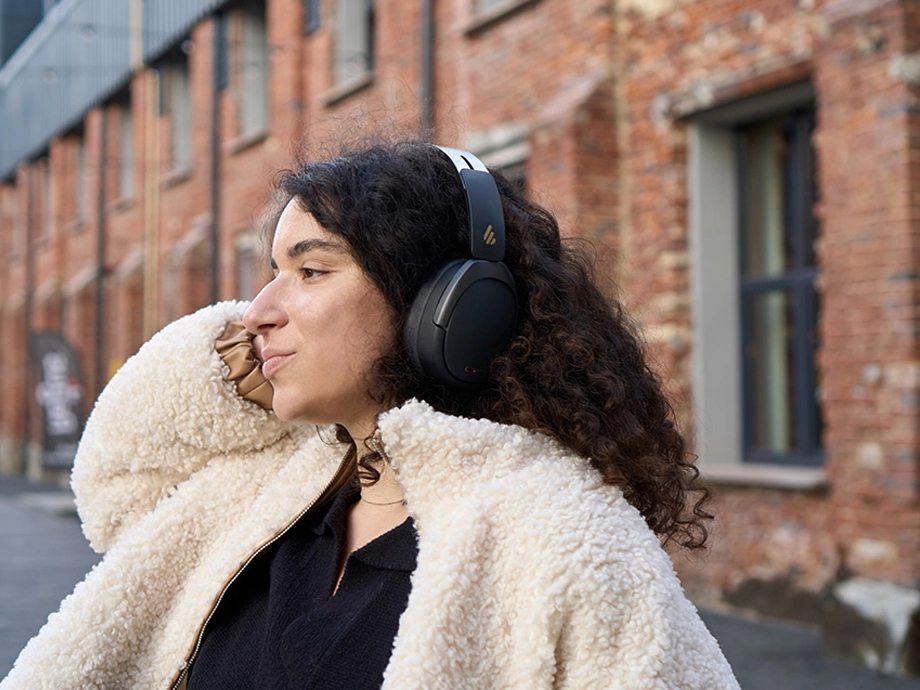 Дівчина з кучерявим волоссям у темних навушниках торкається обличчя. На ній світла куртка.