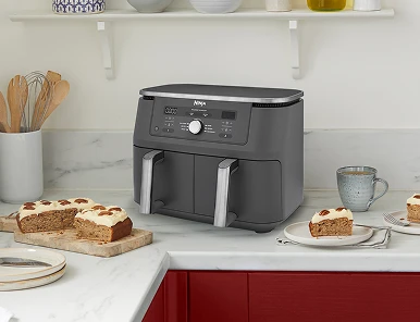 Чорна аерофритюрниця Ninja з двома кошиками стоїть на стільниці у світлій кухні. Поруч з нею лежить торт.