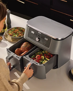 Жінка вставляє або виймає кошики з аерофритюрниці Ninja. У кошиках смажена курка та овочі.