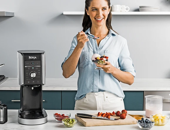 Жінка їсть морозиво, стоячи за кухонною стільницею з різними фруктами та мороженицею Creami Deluxe.