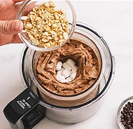 Рука тримає миску з подрібненими горіхами над мискою з шоколадним кремом та зефіром