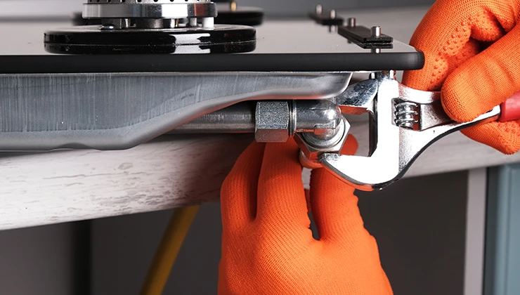 Руки в помаранчевих рукавичках затягують металеве газове з'єднання під чорною пластиною гайковим ключем.