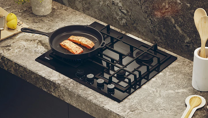 Чорна газова варильна поверхня в кухонному обстановці, на якій стоїть сковорода зі шматочками риби, що смажиться.