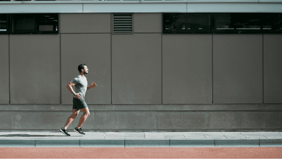 Чоловік біжить тротуаром на тлі бетонної стіни будівлі, одягнений у чорні навушники FreeClip2 та смарт-годинник.