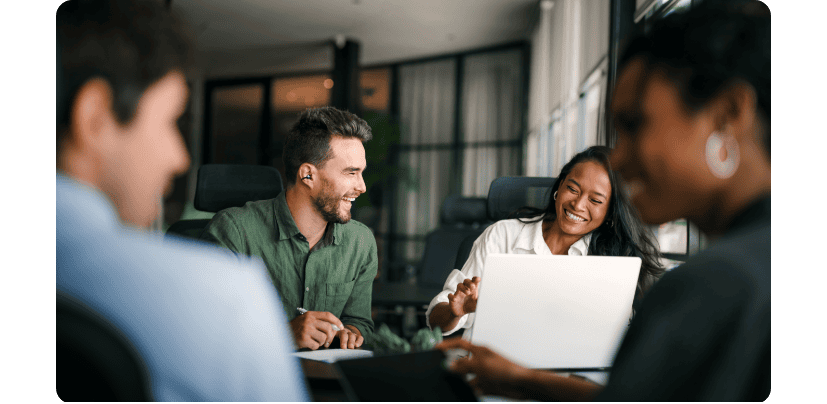 Четверо людей сидять за столом, на якому лежить білий ноутбук, і щасливо посміхаються. У чоловіка у вусі навушники FreeClip 2.