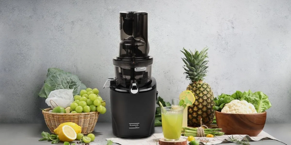 Чорна соковижималка Kuvings Revo830 зі світло-зеленим соком, фруктами та овочами на стільниці.