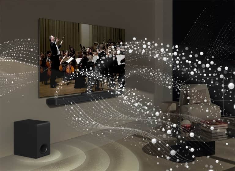 Obraz LG TV i LG Soundbar w salonie. Odtwarzany jest występ muzyczny. Białe krople tworzą fale dźwiękowe, które wystrzeliwują z soundbara i z telewizora w górę i do przodu. Subwoofer tworzy efekt dźwiękowy emitowany z dołu.