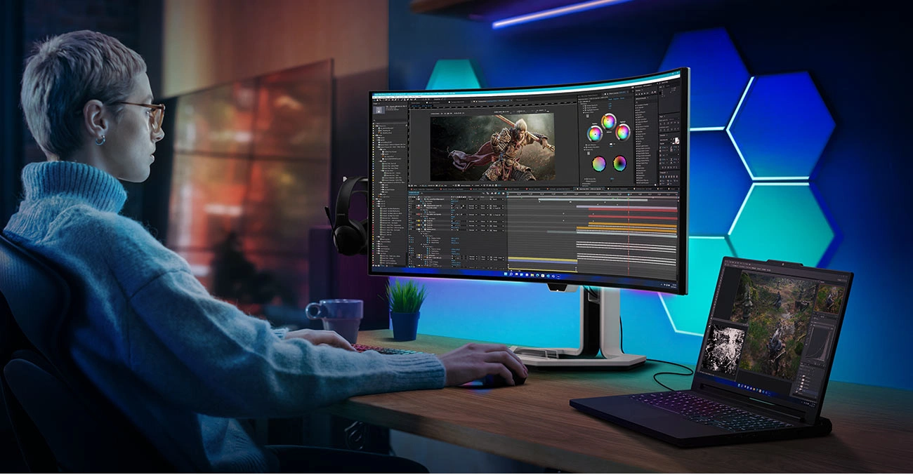 Монітор Lenovo Legion Pro 34WD-10 на столі, перед ним сидить молода людина, поруч — відкритий ноутбук, підключений до монітора.