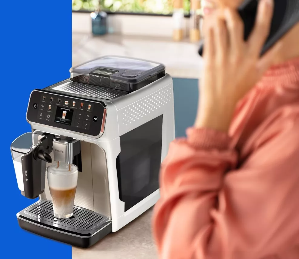 Жінка розмовляє по телефону, а позаду неї видно кавоварку Philips EP5543/80, яка готує каву з щедрою кількістю піни. Жінка розмовляє по телефону, а позаду неї видно кавоварку Philips EP5543/80, яка готує каву з щедрою кількістю піни.