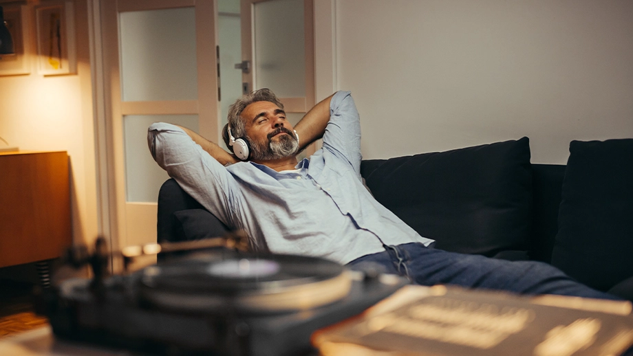 Чоловік слухає музику в бездротових навушниках, лежачи на дивані. На передньому плані видно програвач платівок.