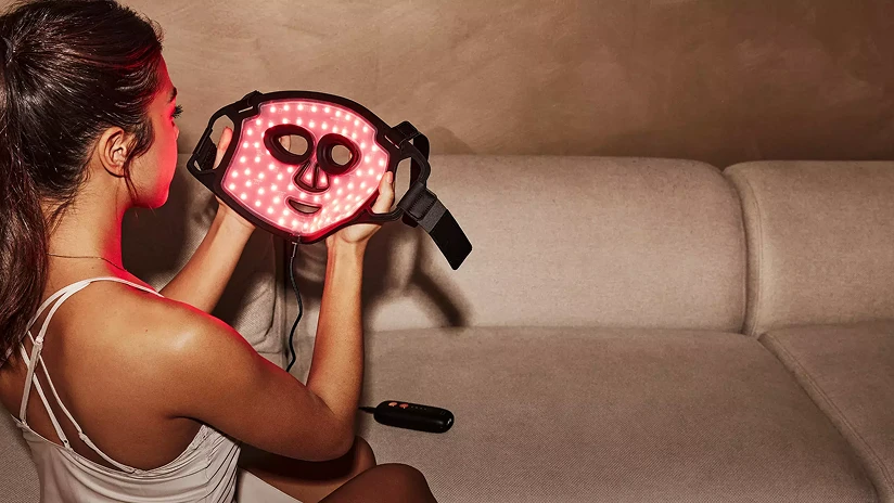 Жінка сидить на дивані, тримаючи чорну маску Silk'n LED (світлодіодна), що світиться інтенсивним червоним світлом.