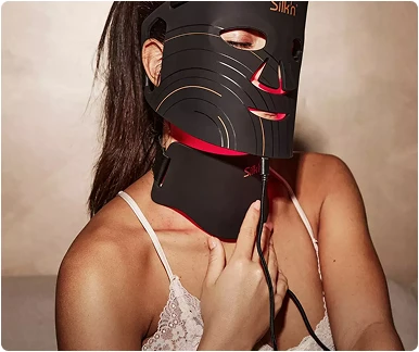 Жінка носить чорну маску Silk'n LED (світлодіодна) на обличчі та шиї, яка випромінює інтенсивне червоне світло.