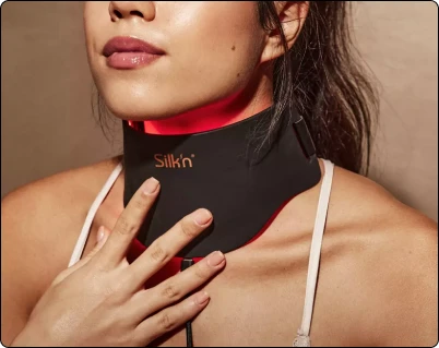 Жінка торкається свого декольте, одягнувши на шию чорний нашийник Silk'n LED (світлодіодний), який випромінює червоне світло.