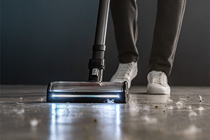 Пилосос Tefal X-Force Flex з увімкненою лампочкою збирає бруд з підлоги, на задньому плані видно ноги людини.