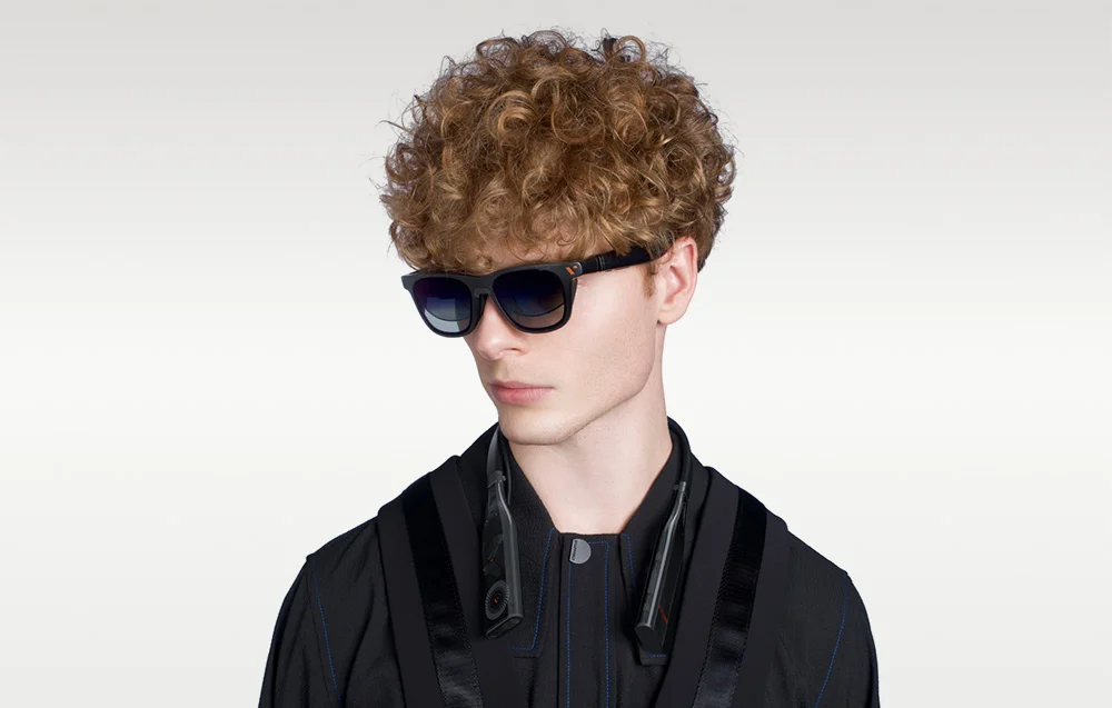 Чоловік у чорному одязі та кучерявому волоссі, в окулярах та з пов'язкою на світлому фоні