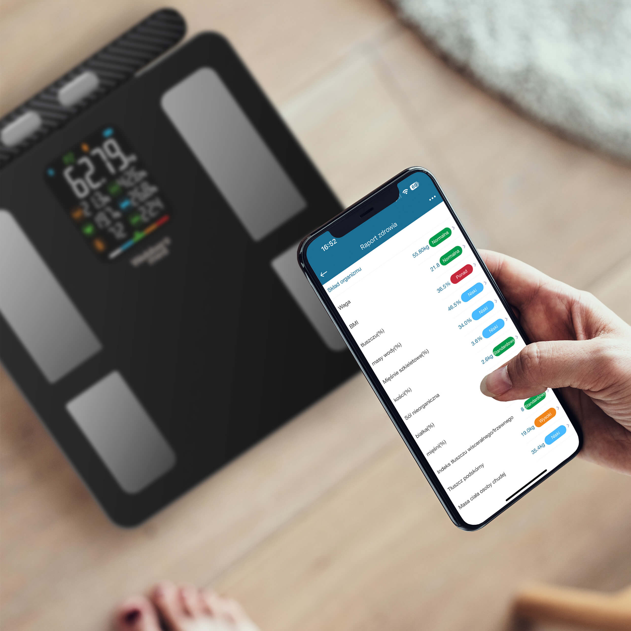 Людина тримає смартфон із фітнес-додатком, а на підлозі поруч лежать чорні електронні ваги з кольоровим дисплеєм.