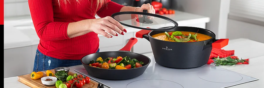 Patelnia i Garnek Obsidian na płycie indukcyjnej. Kobieta unosi pokrywkę patelni, obok deska do krojenia, przyprawy i warzywa