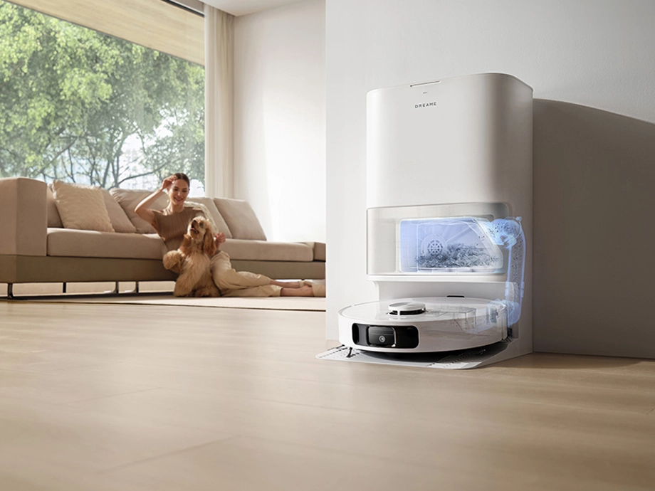 Робот прикреплён к док-станции и опорожняет пылесборник. Вдалеке, в гостиной, видна женщина с собакой.