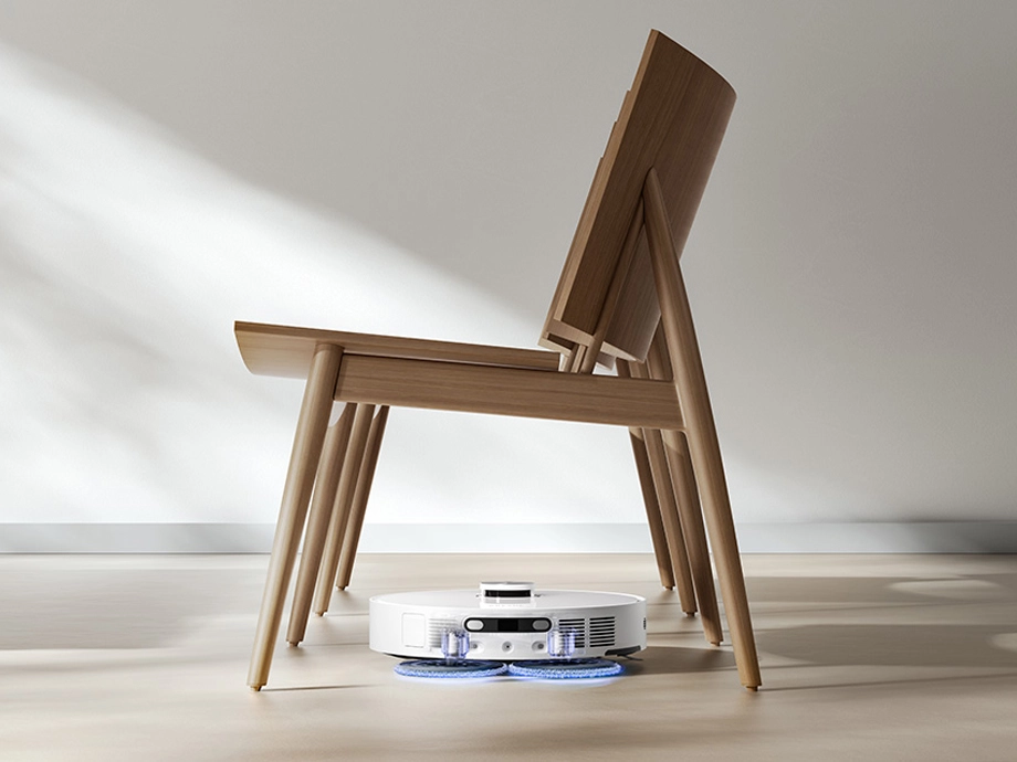 Робот моет светлый пол под деревянным стулом в ярко освещенной комнате.