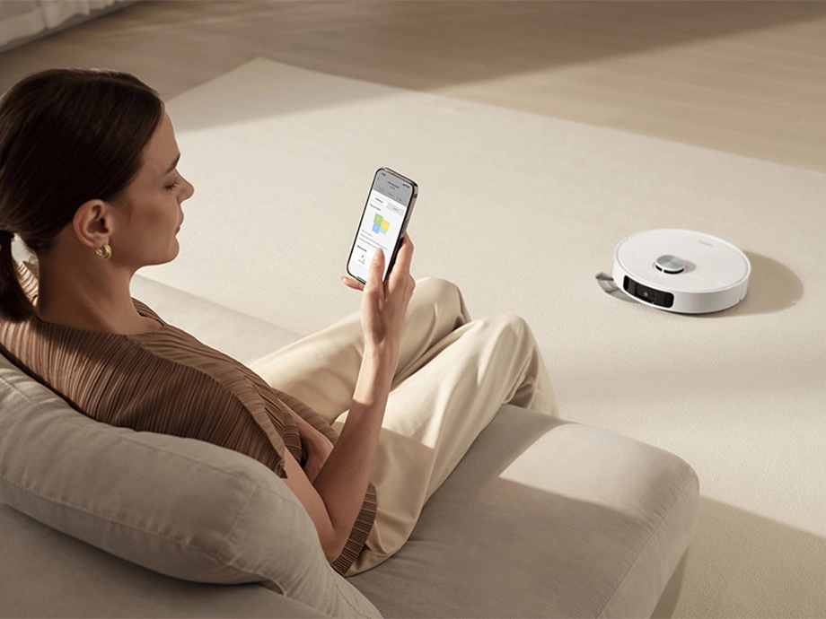 Женщина сидит на диване со смартфоном в руке, а справа робот пылесосит ковер.