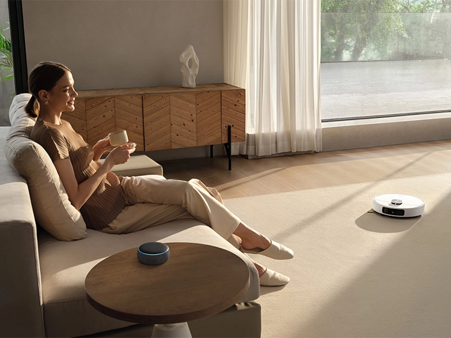 Женщина сидит на диване с чашкой в руке, голосовой помощник лежит на столе, а робот пылесосит светлый ковер.
