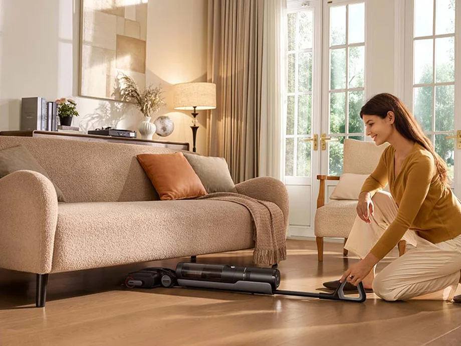 Жінка пилососить простір під диваном, на задньому плані велике вікно.