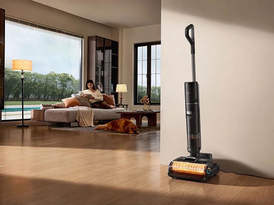 Пилосос стоїть на зарядній станції біля стіни у вітальні, на задньому плані жінка сидить на дивані та читає книгу, а собака лежить на килимі.