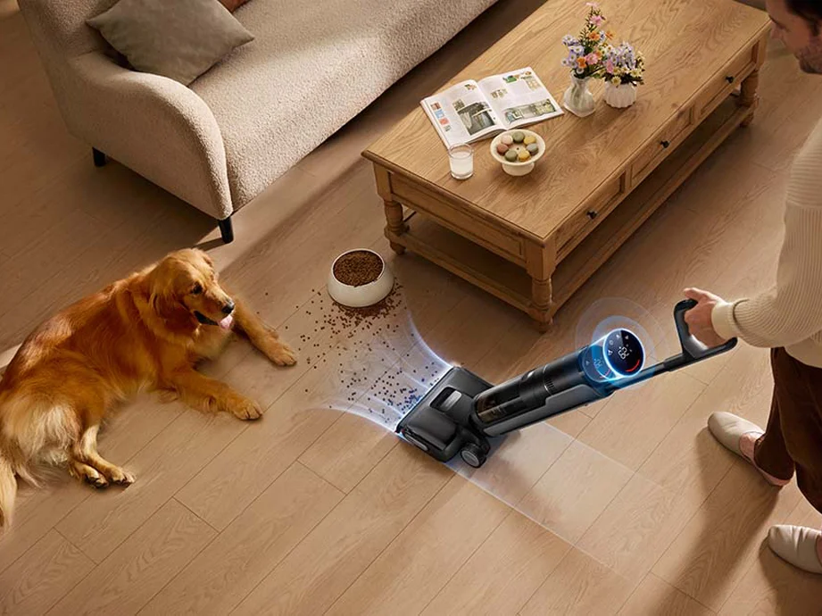 Чоловік пилососить розлитий корм для собак біля журнального столика, поруч лежить золотистий ретривер.