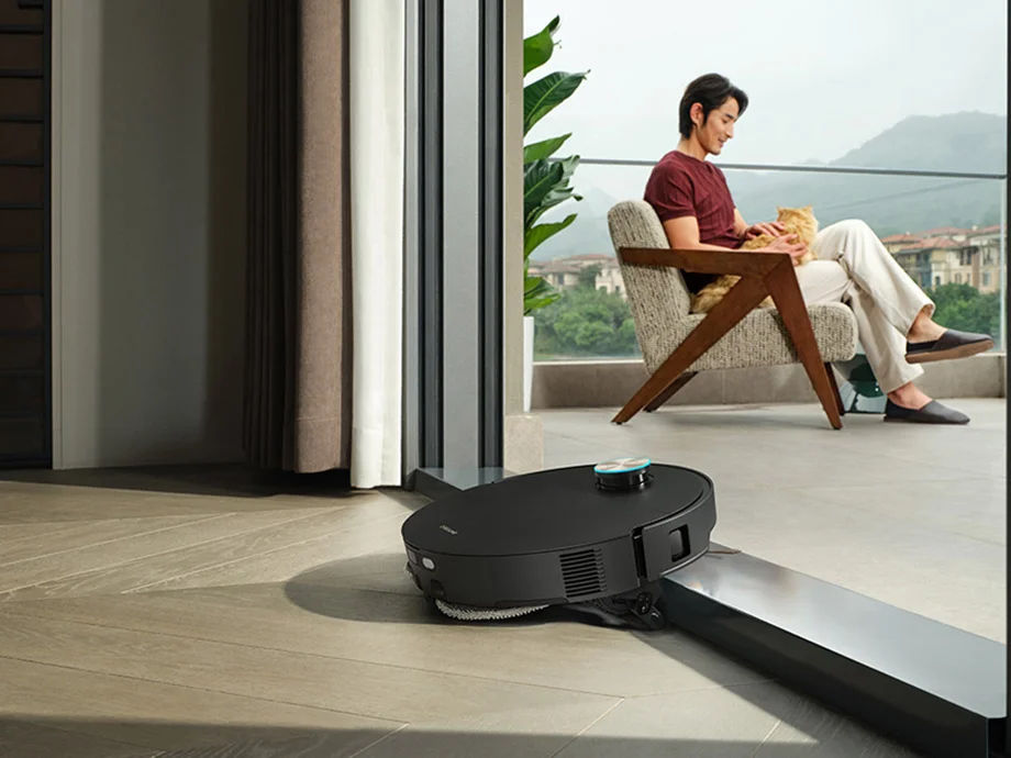 Робот-прибиральник заїжджає на поріг будинку, поки чоловік сидить на балконі.