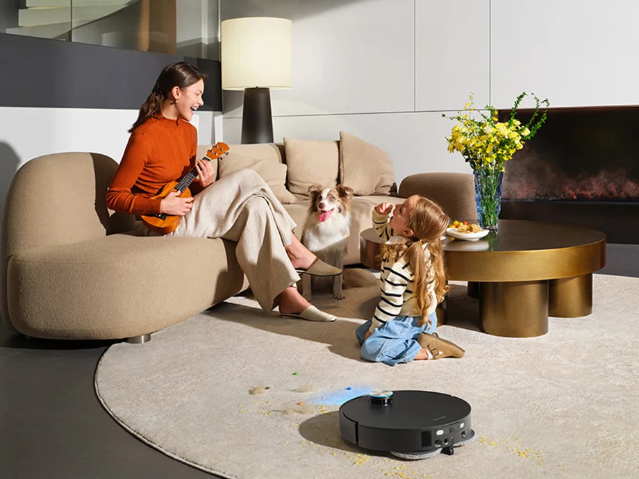 Мати та її донька проводять час разом у світлій, сучасній вітальні, а поруч чорний робот чистить килим.