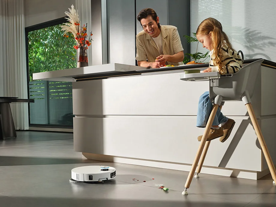 Усміхнений чоловік спирається на кухонний острів, поруч із ним у дитячому стільчику сидить дівчинка, а на підлозі — робот-прибиральник.