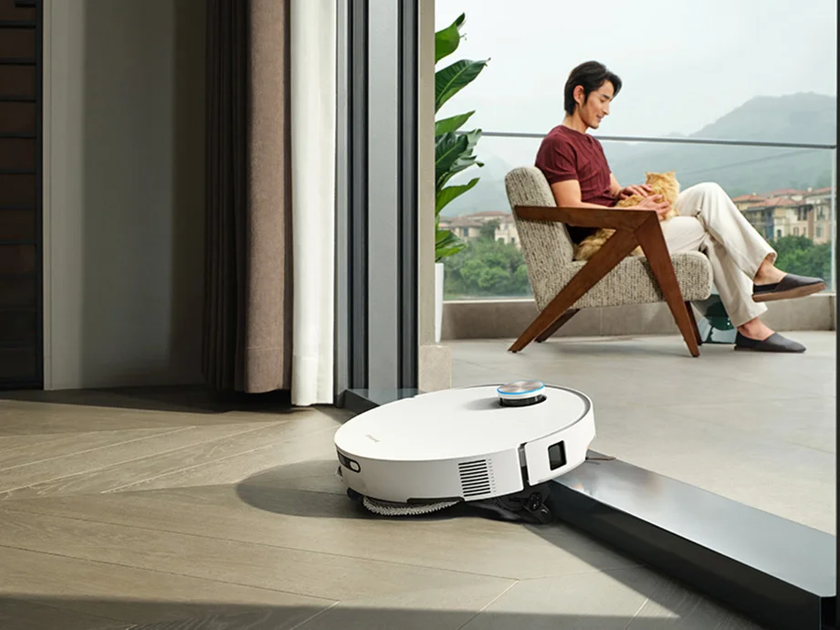 Білий робот-прибиральник заїжджає на поріг, вдалині на балконі сидить чоловік.
