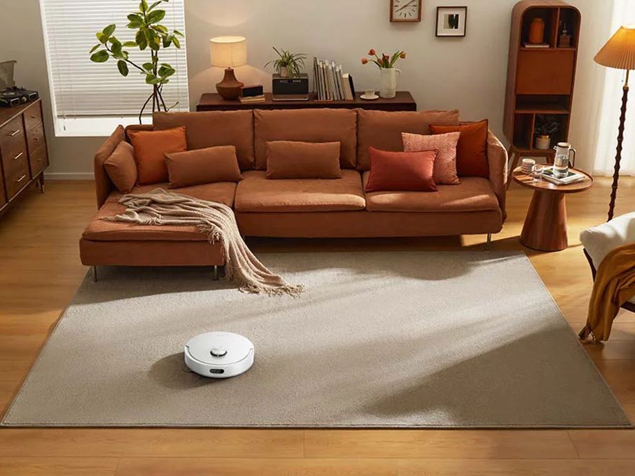 Білий робот-прибиральник на килимі у великій вітальні з коричневими меблями та диваном.