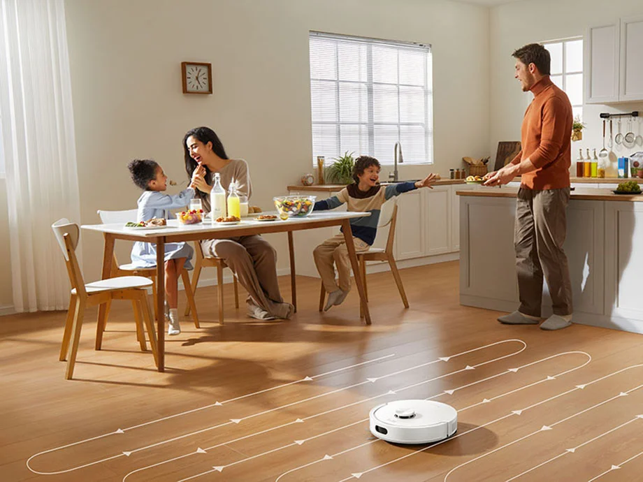 Білий робот-прибиральник пилососить підлогу кухні за заздалегідь визначеним маршрутом, на задньому плані видно сім'ю з дитиною, яка їсть.