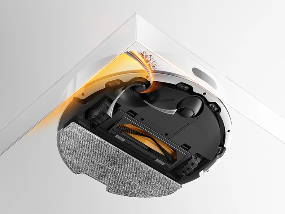 Нижня частина білого робота-пилососа, який видаляє пил уздовж краю стіни за допомогою бічної щітки.