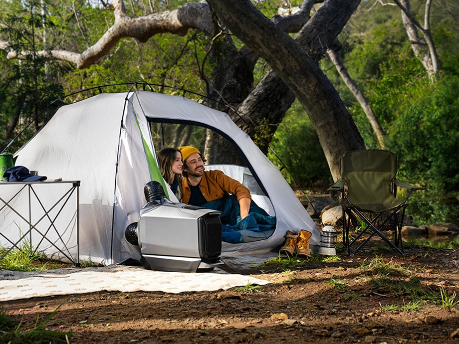 Чоловік і жінка сидять у наметі. Поруч з ними видно кондиціонер, стіл, взуття, лампу та розкладний стілець. На задньому плані дерева.