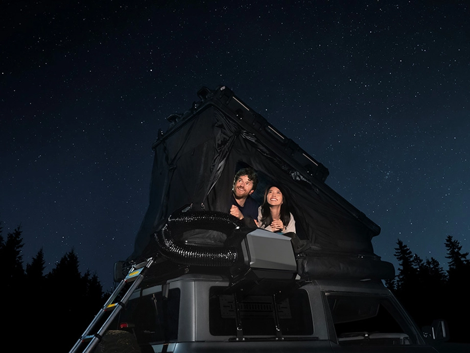 Жінка та чоловік у наметі на даху автомобіля. Поруч видно кондиціонер. Вгорі – зоряне небо.